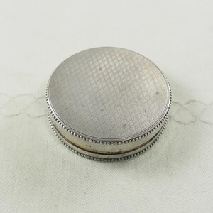 May include: A silver round compact with a textured lid and a beaded edge.