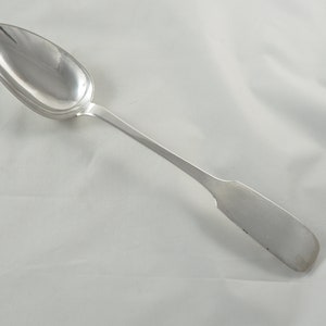 Puede incluir: Una cuchara de plata con un cuenco redondeado y un mango largo y delgado. La cuchara está pulida y brillante.