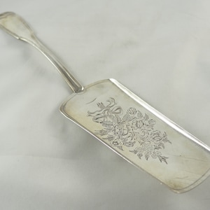 Puede incluir: Un servidor de pastel de plata con un diseño floral. El servidor tiene un mango largo y una hoja rectangular.