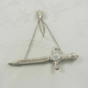 Op de afbeelding: Een zilverkleurige decoratieve zwaardbroche met ingewikkelde details. De broche heeft een zwaardontwerp met een decoratieve handgreep en een ketting verbonden met een veervormig element. De broche is waarschijnlijk van metaal.