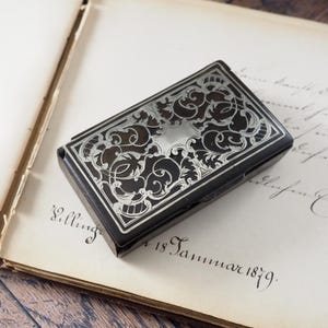 Puede incluir: Una caja rectangular de metal negro con un intrincado patrón de volutas grabado en tono plateado en la tapa. El diseño incluye motivos florales simétricos y un área central vacía en forma de escudo, todo sobre un fondo oscuro.