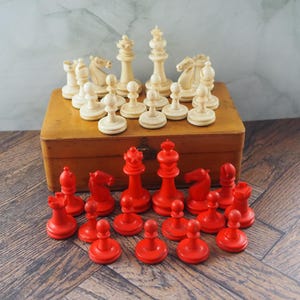 Puede incluir: Un juego de ajedrez vintage con piezas de marfil y rojas. Las piezas de marfil están dispuestas sobre una caja de madera, mientras que las piezas rojas están al frente. El juego incluye peones, torres, caballos, alfiles, una reina y un rey.