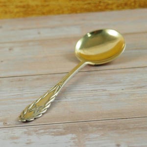 Può includere: Un cucchiaio d'argento con una coppa color oro. Il manico presenta un design decorativo con un motivo floreale. Il cucchiaio è posto su uno sfondo di venature di legno chiaro.