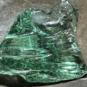 May include: A large piece of green sea glass with a smooth, polished surface. The glass has a translucent quality and shows intricate patterns of lines and swirls.
