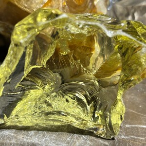 May include: A large, clear, yellow-green piece of glass with a rough, irregular shape. The glass is translucent and has a slightly cloudy appearance.