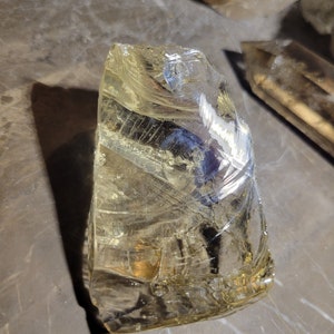 May include: A large, clear, yellow-tinted crystal with a rough, irregular shape. The crystal has a translucent appearance and shows internal striations.