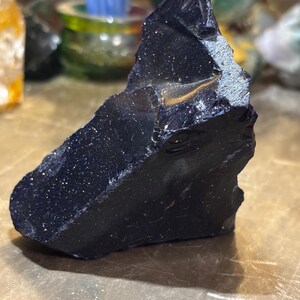 May include: A dark blue, rough-cut gemstone with sparkling inclusions. The stone has a jagged, irregular shape and a polished surface that reflects light. The overall colour is a deep, rich blue with flecks of silver, creating a starry effect. The gemstone is likely a decorative item.