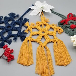 May include: A collection of handmade macrame Christmas ornaments. The ornaments include a navy blue snowflake, a yellow starburst, and a green wreath with red accents. Decorative white berries and red berries are also present.