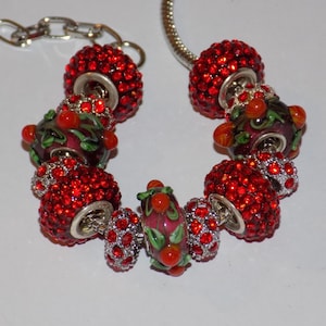 May include: A silver-tone charm bracelet featuring a series of alternating beads. The design includes round beads encrusted with bright red crystals and glass beads decorated with painted green leaves and small red berries on a dark base.