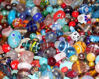 May include: A large collection of assorted glass beads in various shapes, sizes, and colors including blue, red, yellow, green, and white. The beads feature diverse patterns such as stripes, swirls, dots, and translucent finishes, creating a dense pile.