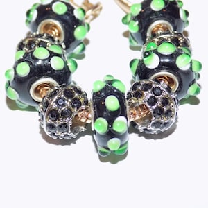 May include: A charm bracelet featuring black glass beads with raised lime green dots, interspersed with metallic spacers encrusted with small black crystals. The beads are strung on a gold-toned metal chain with a visible clasp mechanism at the top.