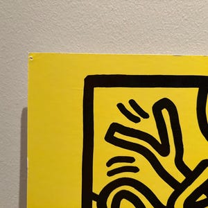 Puede incluir: Una secci&oacute;n de una obra de arte amarillo vibrante con l&iacute;neas negras audaces. La obra presenta una figura humana estilizada, parte de un dise&ntilde;o m&aacute;s amplio. La pieza est&aacute; montada en una pared gris neutra, creando un contraste llamativo.