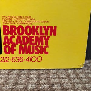Puede incluir: Un cartel amarillo con texto rojo que dice "BROOKLYN ACADEMY OF MUSIC". El cartel incluye el texto "KHARING 1984" y un n&uacute;mero de tel&eacute;fono. El cartel est&aacute; ligeramente da&ntilde;ado en la esquina inferior derecha.