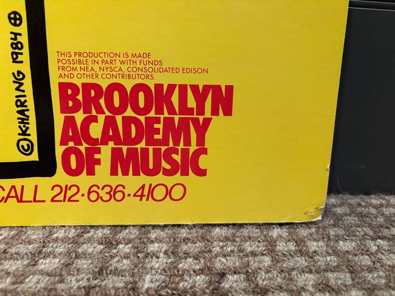 Puede incluir: Un cartel amarillo con texto rojo que dice "BROOKLYN ACADEMY OF MUSIC". El cartel incluye el texto "KHARING 1984" y un n&uacute;mero de tel&eacute;fono. El cartel est&aacute; ligeramente da&ntilde;ado en la esquina inferior derecha.