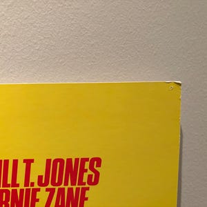 Puede incluir: Una secci&oacute;n de un p&oacute;ster amarillo con el texto "BILL T. JONES ARNIE ZANE" en rojo. El p&oacute;ster est&aacute; contra una pared gris claro. La esquina superior derecha del p&oacute;ster est&aacute; ligeramente da&ntilde;ada.