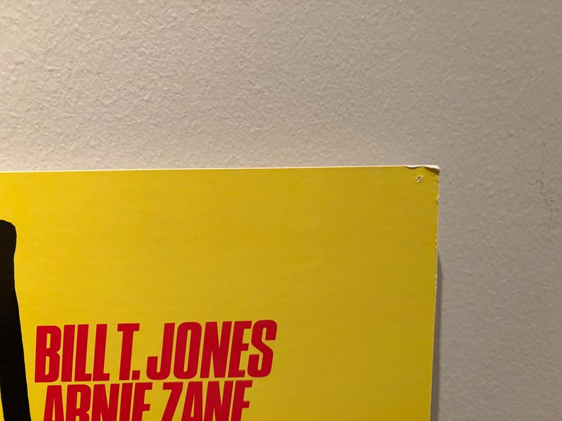 Puede incluir: Una secci&oacute;n de un p&oacute;ster amarillo con el texto "BILL T. JONES ARNIE ZANE" en rojo. El p&oacute;ster est&aacute; contra una pared gris claro. La esquina superior derecha del p&oacute;ster est&aacute; ligeramente da&ntilde;ada.