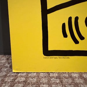 Puede incluir: Una secci&oacute;n de un p&oacute;ster amarillo con elementos gr&aacute;ficos negros. El p&oacute;ster presenta l&iacute;neas y formas negras en negrita, con el texto "Layout and Type: Tom Mennillo" en la parte inferior. El p&oacute;ster est&aacute; sobre una superficie texturizada.