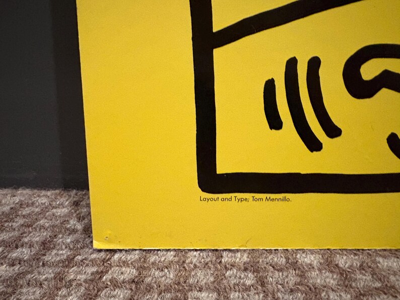 Puede incluir: Una secci&oacute;n de un p&oacute;ster amarillo con elementos gr&aacute;ficos negros. El p&oacute;ster presenta l&iacute;neas y formas negras en negrita, con el texto "Layout and Type: Tom Mennillo" en la parte inferior. El p&oacute;ster est&aacute; sobre una superficie texturizada.