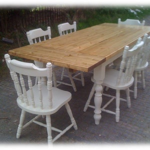 May include: A wooden table with a natural finish and six white chairs. The table has a rectangular top and a single pedestal base. The chairs have a simple design with a slatted back and turned legs.