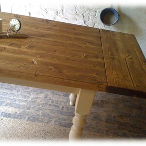 May include: A rustic wooden table with a drop leaf extension. The table top is stained a dark brown color and the legs are painted a light cream color. The table is sitting on a stone floor.