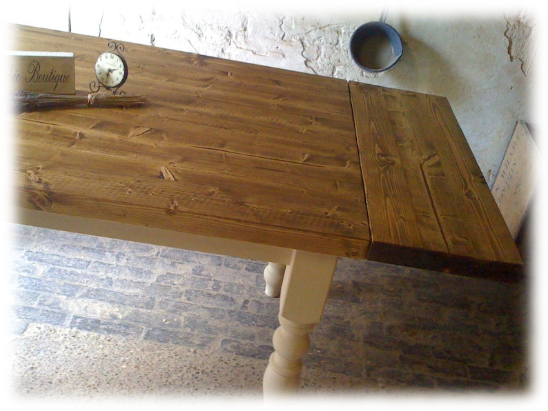 May include: A rustic wooden table with a drop leaf extension. The table top is stained a dark brown color and the legs are painted a light cream color. The table is sitting on a stone floor.