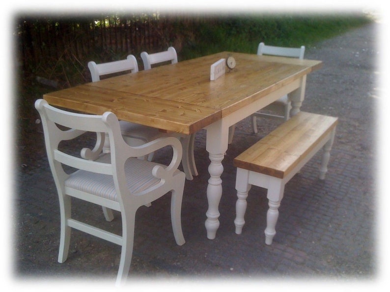 May include: A white farmhouse style dining table with a natural wood top and turned legs. The table is set with four chairs and a bench. The chairs and bench are painted white with a distressed finish.