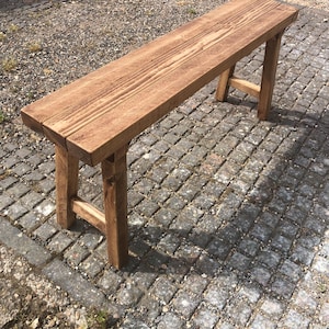 May include: A rectangular wooden bench with a medium brown stained finish and visible wood grain. The bench features a flat seat supported by four sturdy legs connected by horizontal crossbars on each side, creating a stable structure.