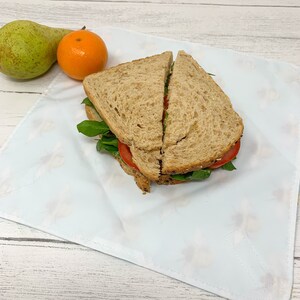 Op de afbeelding: Een witte stoffen wikkel met een bloemenpatroon, gebruikt om een ​​sandwich in te pakken. De sandwich is gemaakt met twee sneetjes volkorenbrood, tomaat, spinazie en een groene spread.