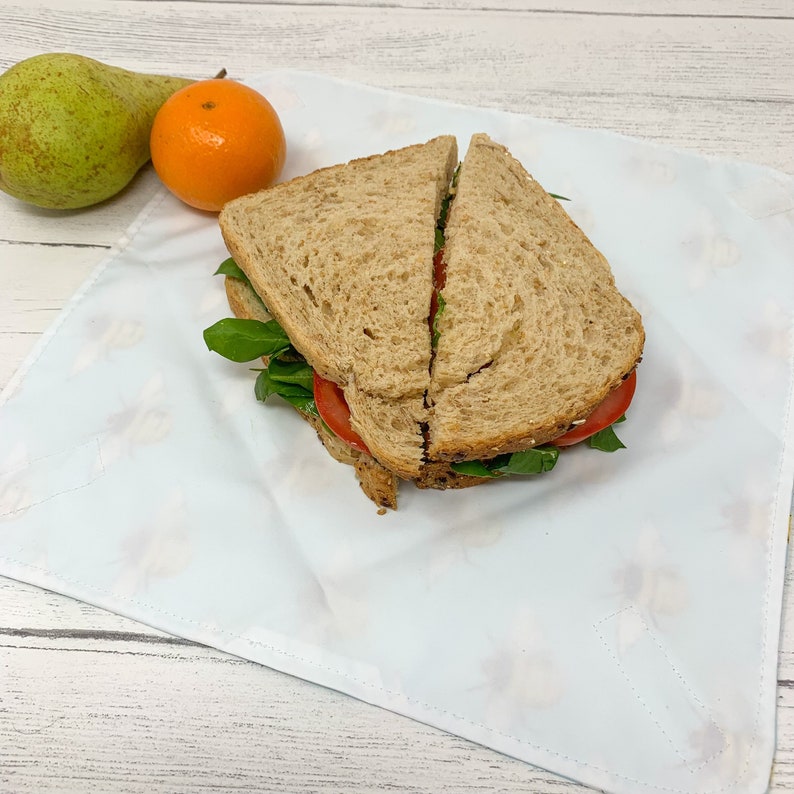 Op de afbeelding: Een witte stoffen wikkel met een bloemenpatroon, gebruikt om een ​​sandwich in te pakken. De sandwich is gemaakt met twee sneetjes volkorenbrood, tomaat, spinazie en een groene spread.