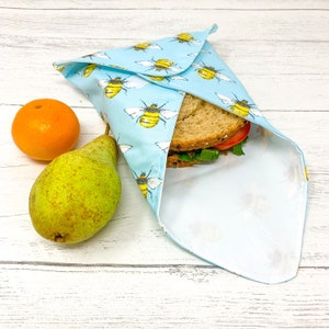 Op de afbeelding: Een blauwe en witte herbruikbare voedselverpakking met een bijenpatroon is gevouwen rond een sandwich. Een peer en een sinaasappel liggen op een wit houten oppervlak.