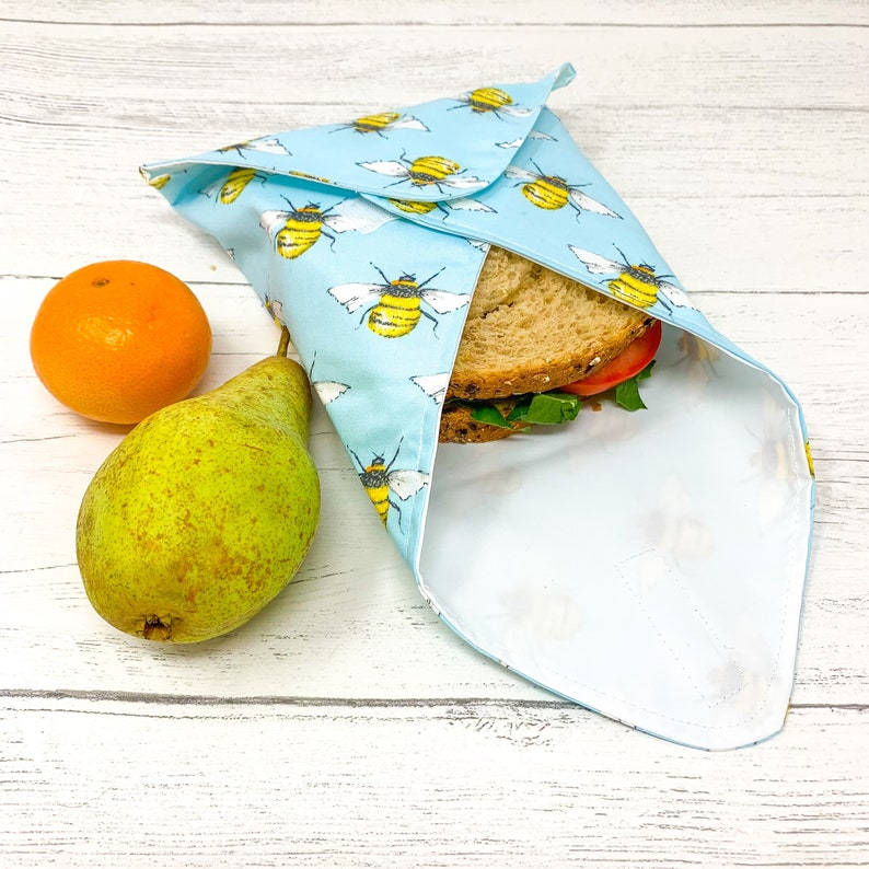 Op de afbeelding: Een blauwe en witte herbruikbare voedselverpakking met een bijenpatroon is gevouwen rond een sandwich. Een peer en een sinaasappel liggen op een wit houten oppervlak.