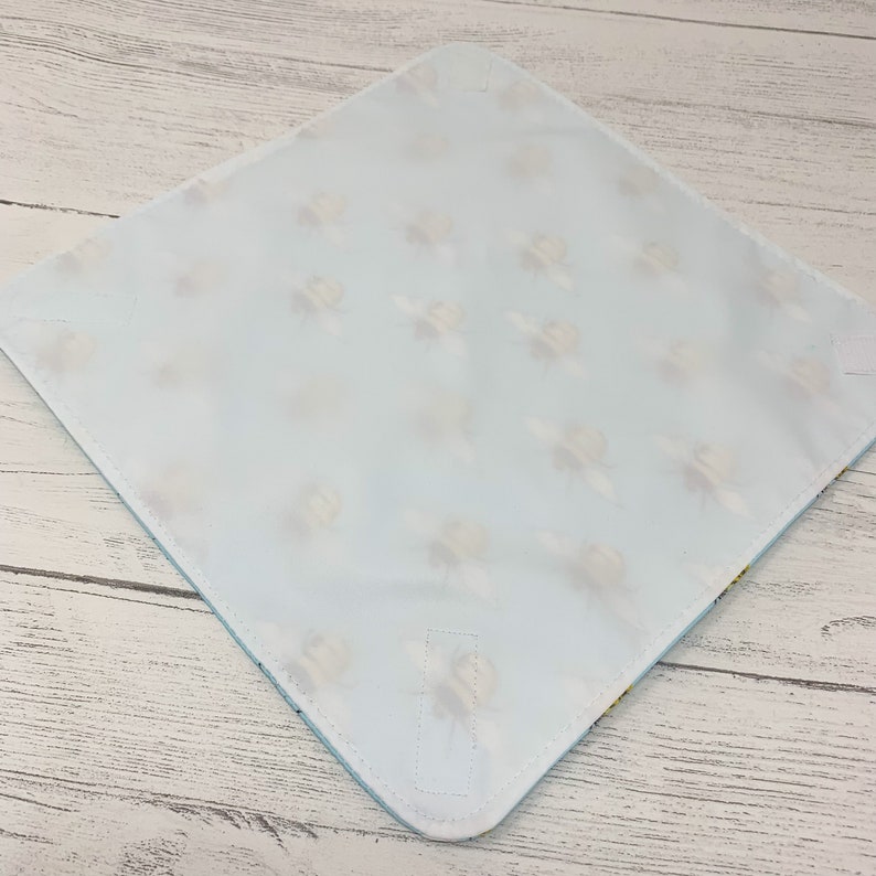 Op de afbeelding: Een lichtblauwe baby spuugdoek met een wit en geel bijenpatroon. De doek heeft een vastgenaaide flap aan de ene kant.