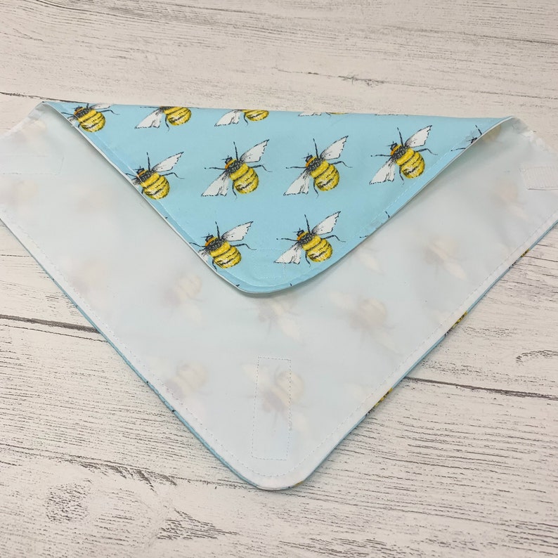 Op de afbeelding: Een lichtblauwe bandana met een witte rand en een patroon van gele en zwarte bijen. De bandana is dubbelgevouwen en het patroon is aan beide kanten zichtbaar.