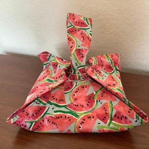 May include: A fabric bag featuring a watercolor-style watermelon pattern with red fruit, black seeds, and green rinds on a light gray background. The bag is constructed with a single handle loop secured by a circular metal ring at the top center.