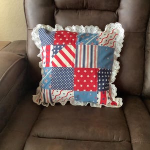 May include: A square patchwork throw pillow featuring a variety of red, white, and blue fabric patterns including stars, stripes, and dots. The pillow is finished with a decorative white eyelet lace ruffle trim along all four edges.