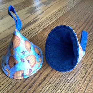 May include: Two fabric pot holders with a blue and yellow bee and honeycomb pattern. One is upright, showing the exterior design and a blue loop. The other is lying on its side, revealing a blue interior.