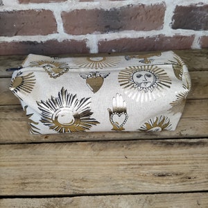 May include: A white canvas cosmetic bag with a gold zipper and a repeating pattern of suns, hearts, and hands. The bag is on a wooden surface.