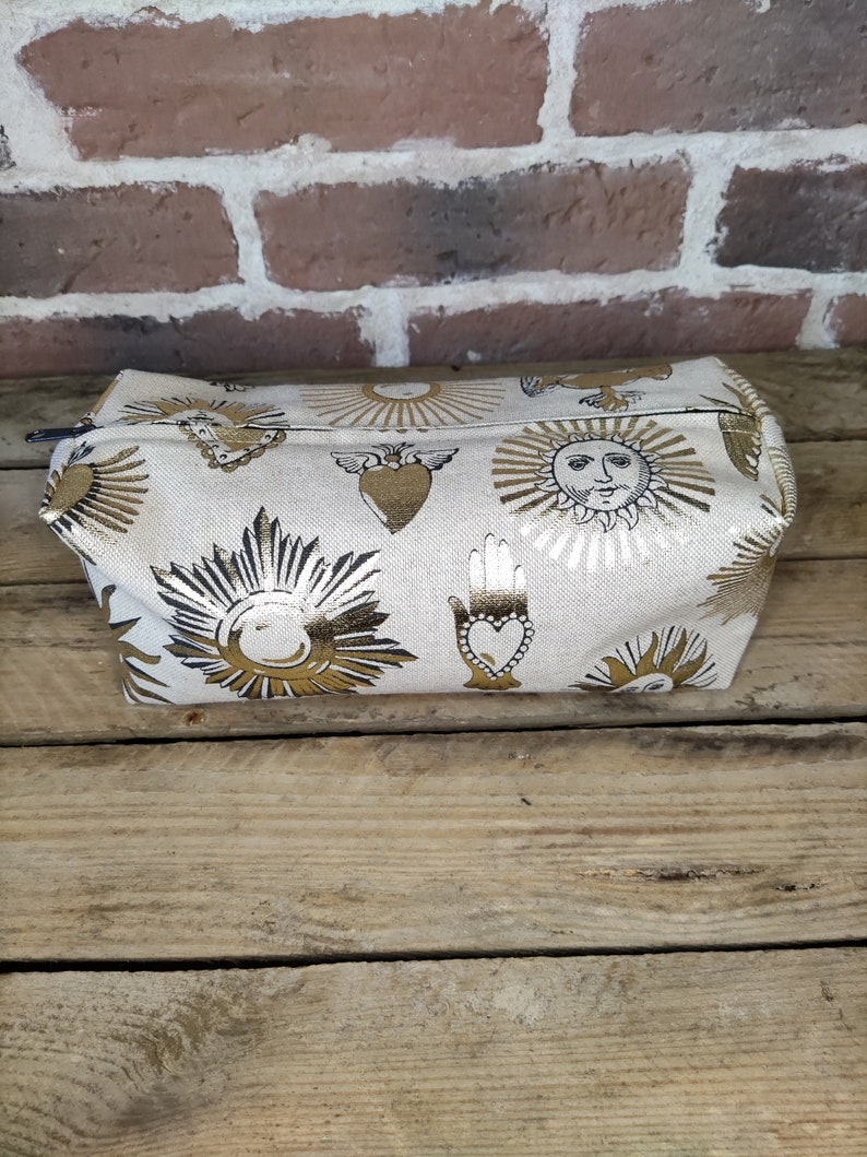 May include: A white canvas cosmetic bag with a gold zipper and a repeating pattern of suns, hearts, and hands. The bag is on a wooden surface.