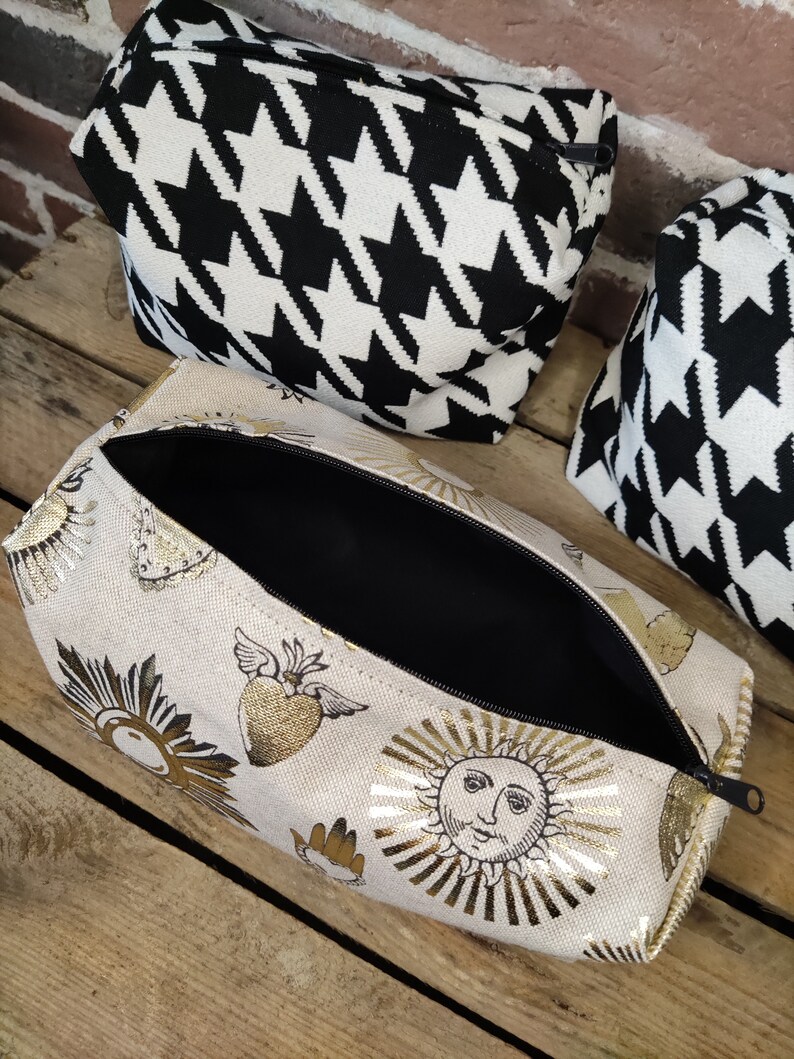 May include: A beige zippered pouch with a gold foil design of suns, hearts, and wings. The pouch is open and the inside is black.