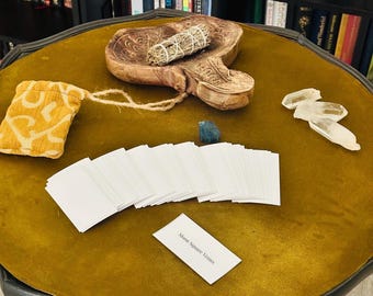 May include: A yellow velvet table top with a stack of white cards, a wooden carving, a small yellow pouch, a bundle of dried sage, and two clear quartz crystals. The card on top of the stack reads "Moon Square Venus".
