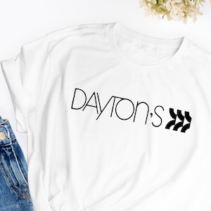 Może przedstawiać: Biała koszulka z czarnym napisem "DAYTON'S" i czarnym geometrycznym wzorem.
