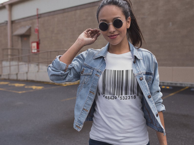 May include: A white t-shirt with a black barcode design and the numbers 9788420532318 printed below. The person is wearing a light blue denim jacket and round sunglasses.