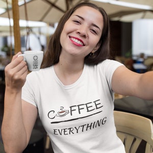 以下が含まれることがあります： コーヒーカップの小さなグラフィックと「COFFEE EVERYTHING」というテキストが書かれた白いTシャツ。小さな白いカップを持った人が着ています。