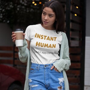 Könnte beinhalten: Weißes T-Shirt mit dem Text "INSTANT HUMAN (JUST ADD COFFEE)" in goldener Schrift. Die Person trägt eine blaue Jeans-Shorts und einen hellgrünen Cardigan.