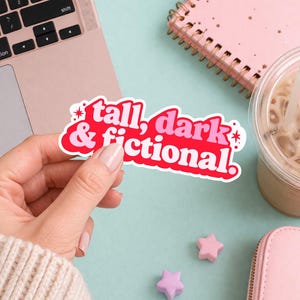 Op de afbeelding: Een sticker met de tekst "tall, dark & fictional" in witte en roze letters op een rode achtergrond. De sticker wordt door een persoon vastgehouden. Andere items op het lichtblauwe oppervlak zijn een laptop, een roze notitieboek en een drankje.