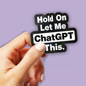 Op de afbeelding: Zwarte sticker met witte tekst: "Hold On Let Me ChatGPT This." De sticker wordt door een hand vastgehouden tegen een lichtpaarse achtergrond. De tekst is vetgedrukt en sans-serif.