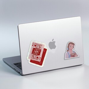 May include: A laptop with stickers of playing cards and a cartoon character. One sticker is a red playing card with the text "QUEEN OF BOOKS". Another sticker is a cartoon of a woman holding a book with the word "SMUT".