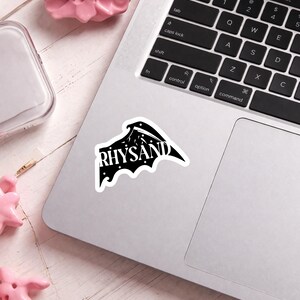 Puede incluir: Una pegatina en blanco y negro con la palabra "RHYSAND" en una fuente estilizada. La pegatina tiene forma de ala de murci&eacute;lago con estrellas y una luna creciente. Est&aacute; colocada sobre un port&aacute;til plateado.