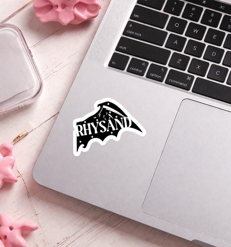 Puede incluir: Una pegatina en blanco y negro con la palabra "RHYSAND" en una fuente estilizada. La pegatina tiene forma de ala de murci&eacute;lago con estrellas y una luna creciente. Est&aacute; colocada sobre un port&aacute;til plateado.