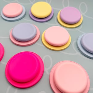 以下が含まれることがあります： パステルカラーの丸いシリコン製の蓋のコレクション。蓋は積み重ねられており、小さく明るい色の蓋が、より大きく暗い色のベースの上に配置されています。ピンク、黄色、紫、青などの色があります。